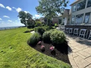 A landscaped backyard with a mulched garden bed featuring ornamental grasses, shrubs, and colorful flowers, next to a paver patio with bar stools and outdoor seating. The yard overlooks a waterfront view with a metal fence, lush green grass, and a bright blue sky with scattered clouds.