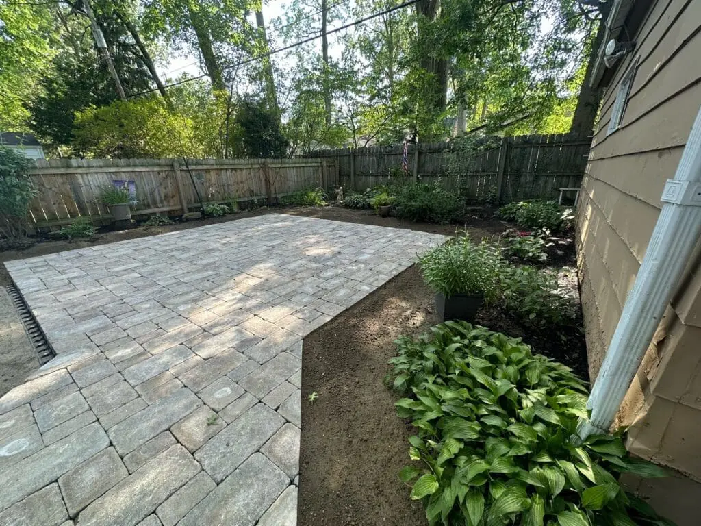 A newly installed stone patio surrounded by freshly mulched flower beds with green plants and shrubs. The area is enclosed by a wooden fence with tall trees in the background, creating a serene backyard environment.