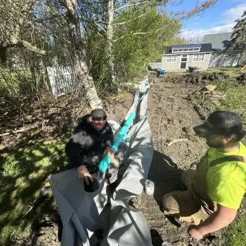Installing Backyard Drainage System Two workers installing a drainage system with a blue pipe wrapped in fabric liner in a backyard. The trench runs alongside trees and shrubs, with a small shed and white picket fence visible in the background under a bright blue sky.