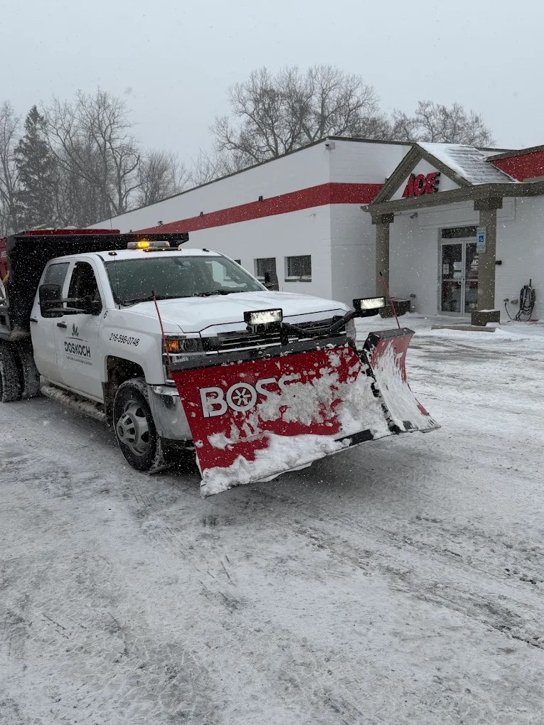 Commercial snow plow truck clearing snow from a parking lot during winter storm conditions in front of a retail building.