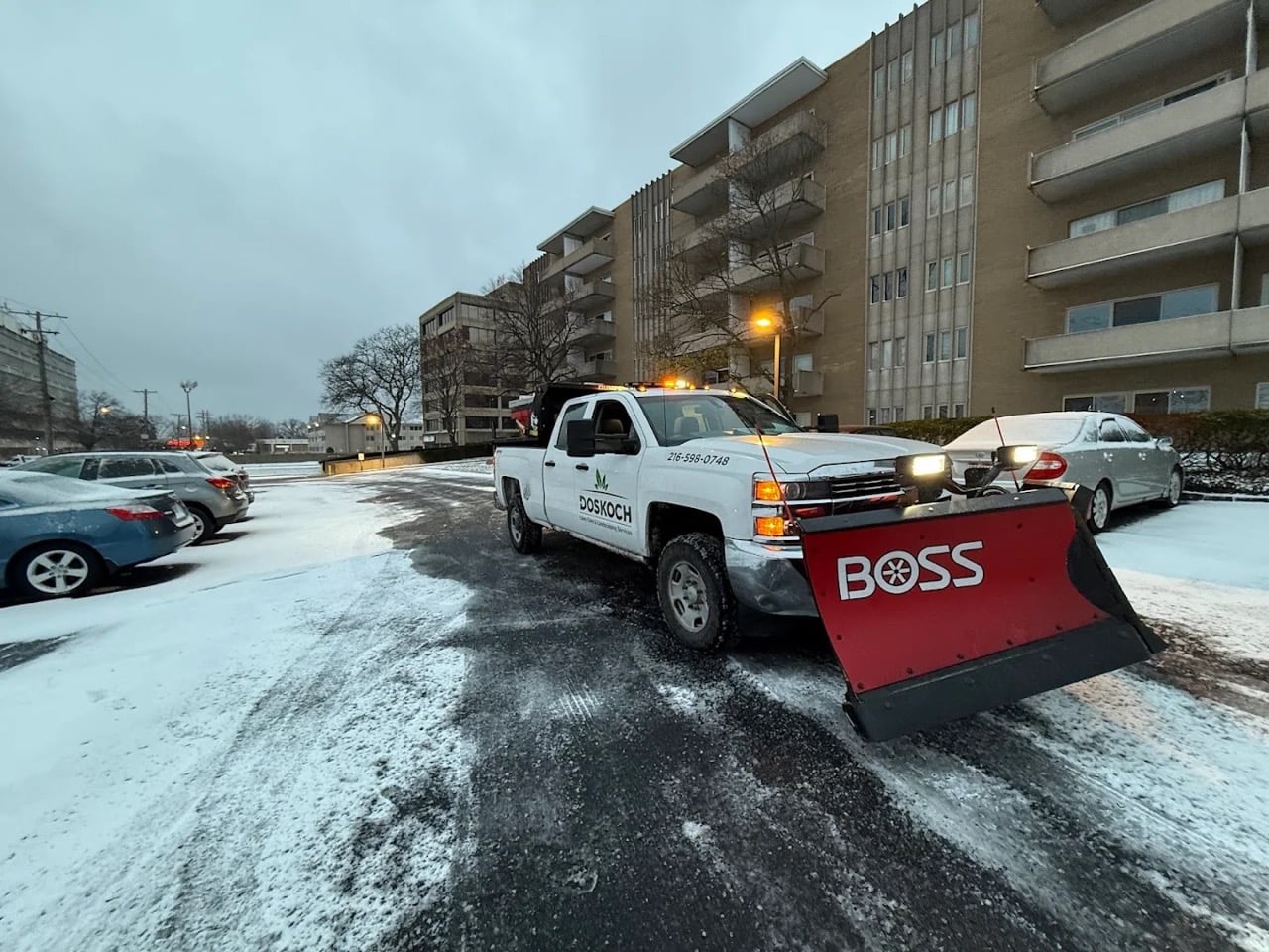 Snow plow truck clearing a commercial parking lot during early morning snowfall near an apartment building.