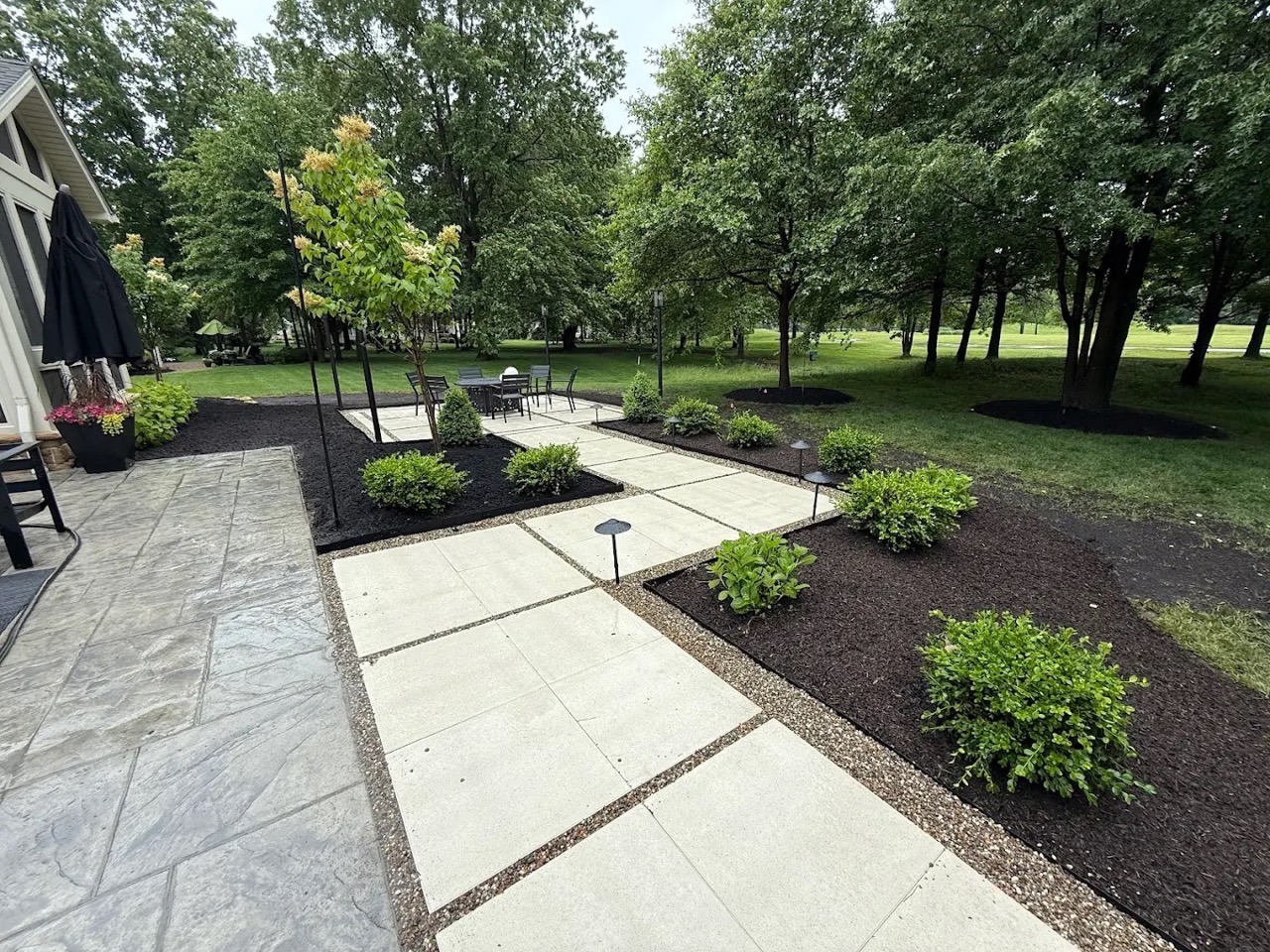 Backyard hardscaping with a paver patio and stone walkway, bordered by fresh mulch beds, trimmed shrubs, and landscape lighting in a wooded residential yard.