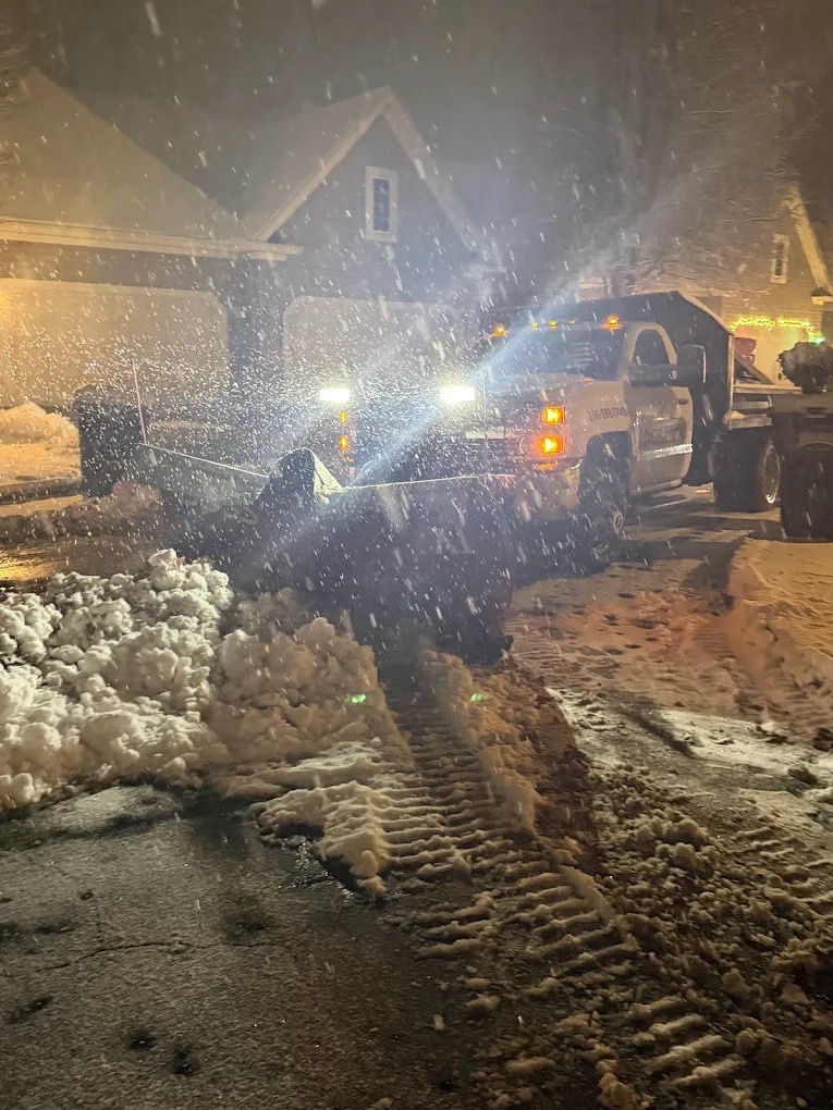 Snow plow truck clearing a residential driveway at night during heavy snowfall