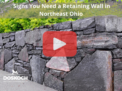 Rock retaining wall. Red play buttom overlay and Doskoch logo. Words: Signs You Need a Retaining Wall in Northeast Ohio