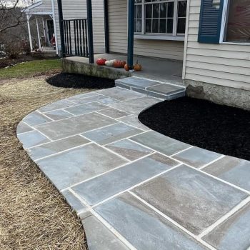 Blue Stone Walkway with Mulched Borders A newly installed blue stone walkway leading to the front steps of a house with beige siding and blue shutters. The pathway is bordered by freshly mulched garden beds and straw-covered soil for new grass seeding. Small decorative pumpkins are placed near the steps, and the yard features bare trees and a quiet suburban setting.